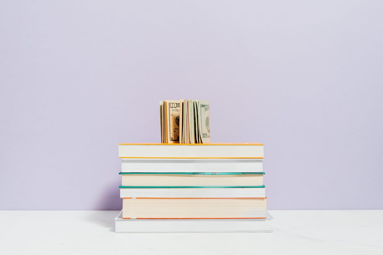 Stack of books with a stack of US dollar bills on top against a pastel background.